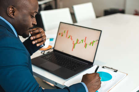 dark-skinned african american businessman student work at laptop with stock market trading terminal....