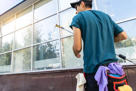 A young male worker of a professional cleaning service in overalls washes the glass of the windows...