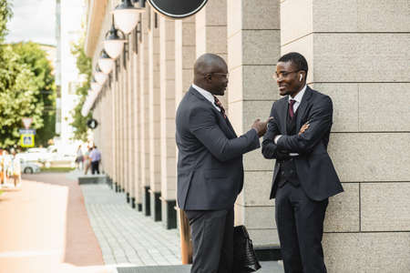 A couple of black African American businessmen friends in suits are talking while standing on the street. Outdoor business meetingの写真素材