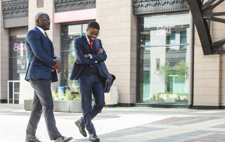 Two dark-skinned african american businessmen in suits and glasses walk outdoors in the city and discuss businessの写真素材