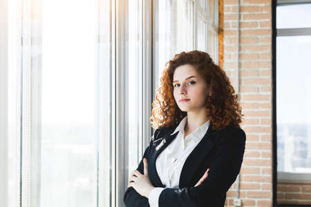 Portrait of a beautiful successful young business woman with red curly hair in the office interior stands near the windowの写真素材