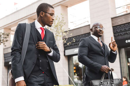 Two dark-skinned african american businessmen in suits and glasses walk outdoors in the city and discuss businessの写真素材
