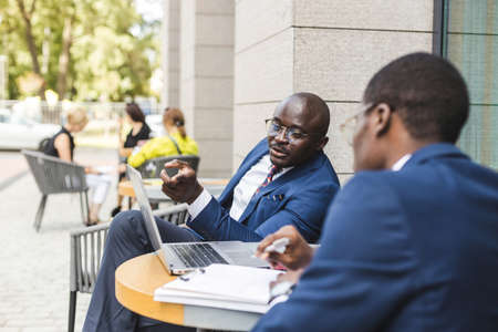 Two dark-skinned African American businessmen in suits and glasses with briefcases sit at a table in an outdoor city cafe.の写真素材