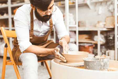 A young male potter is engaged in craft in his workshop on a potters wheel and makes a clay productの写真素材
