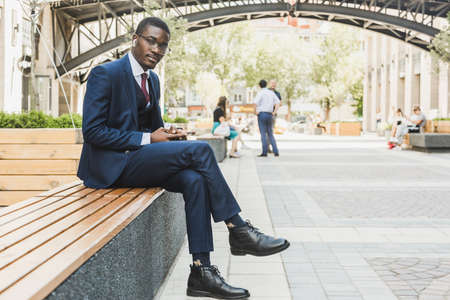 Portrait of a black African American businessman in a suit, glasses and headphones sits on a bench in a city square outdoorsの写真素材