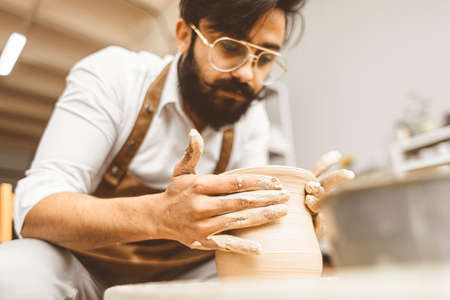 A young male potter is engaged in craft in his workshop on a potters wheel and makes a clay productの写真素材