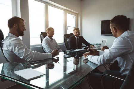 A group of company employees sits at a table in a meeting room. A team of young businessmen working and communicating together in an office. Corporate business team and manager in a meetingの写真素材