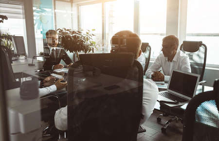 A group of company employees sits at a table in a meeting room. A team of young businessmen working and communicating together in an office. Corporate business team and manager in a meetingの写真素材