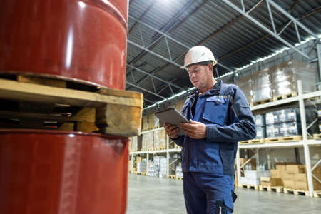 A worker in overalls and a helmet in a warehouse inspects products stored in barrels. Calculation and verification of reserves in an oil company, solving logistics problemsの写真素材