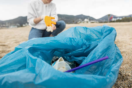 A woman in gloves with a special blue bag picks up garbage among the sand along the coast. The...