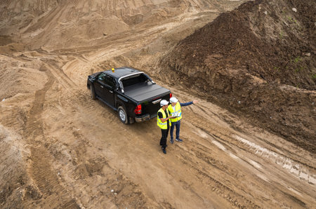 Two male engineers in overalls and white helmets stand near a special vehicle against the background of the construction of a road or facility and working equipment.の写真素材