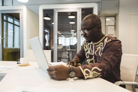 A black African American businessman in a national ethnic business suit works at a laptop. Analytics and work onlineの写真素材