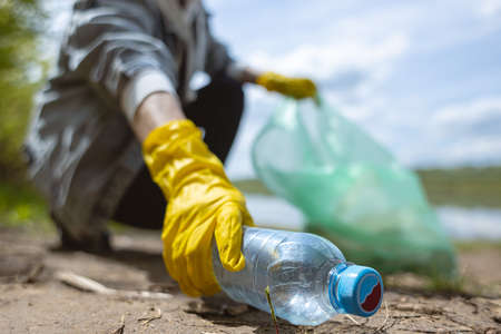 A young woman in yellow gloves collects abandoned garbage in a black bag in the forest. Plastic pollution and environmental protectionの写真素材