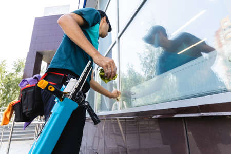 A young male worker of a professional cleaning service in overalls washes the glass of the windows of the facade of the building. Clean showcases on requestの写真素材