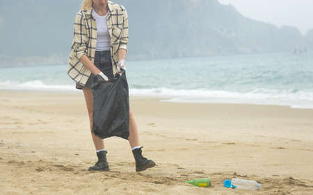 A young woman collects discarded garbage from the beach into a black bag. plastic environmental...