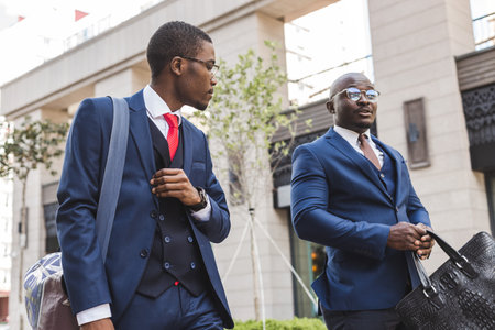 Two dark-skinned african american businessmen in suits and glasses walk outdoors in the city and discuss businessの写真素材