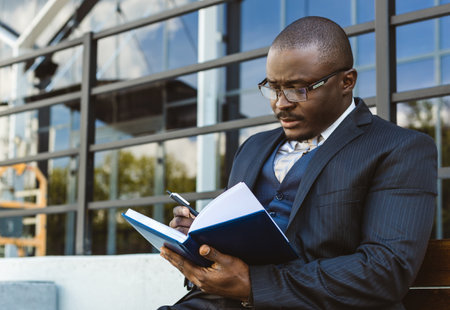 A dark-skinned businessman in a suit reads an outdoor notebook while sitting on a bench against the backdrop of city buildingsの写真素材