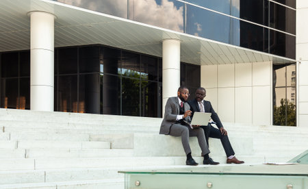 Friendly meeting of business partners outdoors. Two dark-skinned men in suits sit on the steps of a city building with a notebook and have a conversation. Working break. Support and collaborationの写真素材
