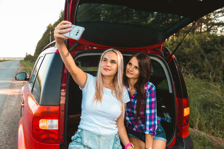 Young pretty women friends travel by car. Summer holidays and active recreation. Positive emotions from the adventure, Girls take a selfieの写真素材