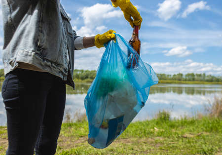 A young woman in yellow gloves collects abandoned garbage in a black bag in the forest. Plastic pollution and environmental protectionの写真素材