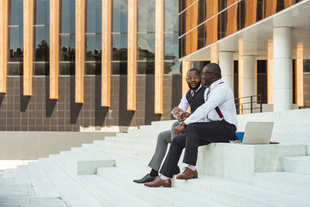 Two African American businessmen in suits communicate. Friends and business partners sit on the concrete steps of the city stairs against the backdrop of a modern skyscraperの写真素材