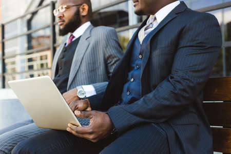 Business meeting of friends outdoors. Two dark-skinned men in suits are sitting on a bench near a...
