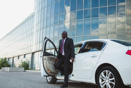 Portrait of a handsome successful African American businessman in a suit getting out of his personal car against the backdrop of a glass building in the city centerの写真素材