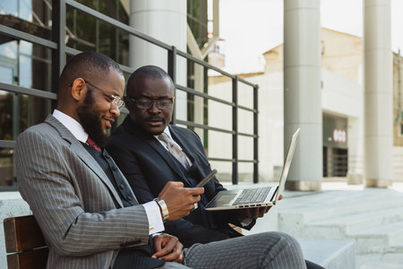 Business meeting of friends outdoors. Two dark-skinned men in suits are sitting on a bench near a city building with a laptop and talking. Partnership and corporate relationsの写真素材