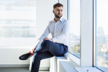 Portrait of a business young successful man in a modern office sitting by the window against the backdrop of the cityの写真素材