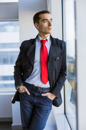 Portrait of a business young successful man in a modern office standing by the window against the...