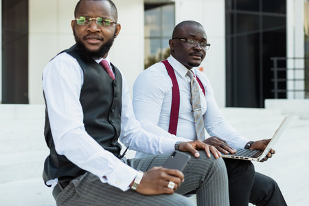 Business meeting outdoors. Two dark-skinned men in suits sit on the steps of a city building with a...