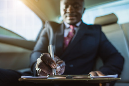 Business concept of a handsome dark-skinned man in a suit and glasses in the back seat of a luxury car signing documents and making work notesの写真素材