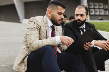 Business people outdoor meeting. A company of male businessmen in suits are sitting on the steps of the stairs. Working break. Teamwork and brainstorming. Successful teamworkの写真素材