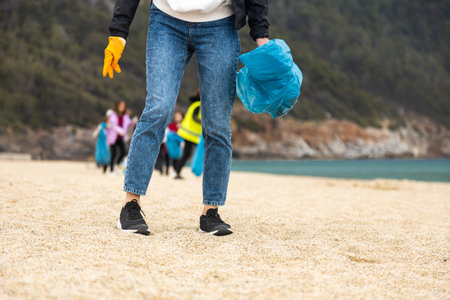 A woman in gloves with a special blue bag picks up garbage among the sand along the coast. The problem of environmental pollution. Cleaning up trash on the beachの写真素材