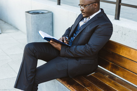 A dark-skinned businessman in a suit reads an outdoor notebook while sitting on a bench against the backdrop of city buildingsの写真素材