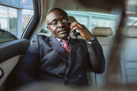 Portrait of a handsome dark-skinned businessman in a suit and glasses in the back seat of a luxury car talking on the phoneの写真素材