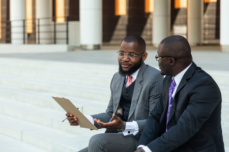 Friendly meeting of business partners outdoors. Two dark-skinned men in suits sit on the steps of a city building with a notebook and have a conversation. Working break. Support and collaborationの写真素材