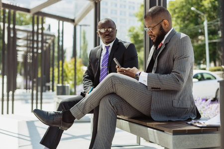 Two dark-skinned businessmen in suits communicate. Friends and business partners are sitting on a bench in the city against the backdrop of a glass buildingの写真素材