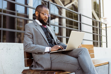 A dark-skinned man in a business suit is using a laptop outdoors. Digital education and remote workの写真素材