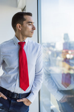 Portrait of a business young successful man in a modern office standing by the window against the backdrop of the cityの写真素材