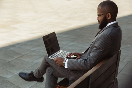A dark-skinned man in a business suit is using a laptop outdoors. Working break. Online learning and education. Distance courses and communication. Copy space and mock upの写真素材