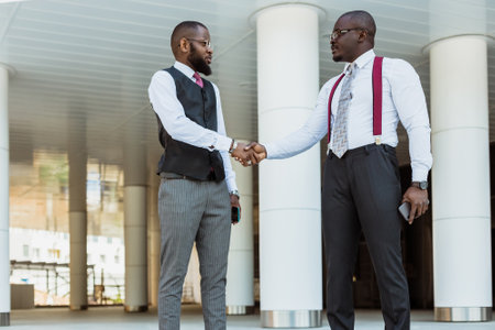 Greeting and handshake of two afro american businessmen partner against the backdrop of a modern building exterior. Friendly meeting outdoorsの写真素材