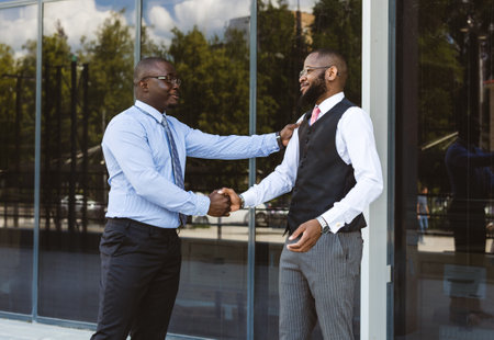 Greeting and handshake of two afro american businessmen partner against the backdrop of a modern building exterior. Friendly meeting outdoorsの写真素材