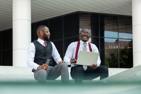 Business meeting outdoors. Two dark-skinned men in suits sit on the steps of a city building with a laptop and have a conversation. Friendly support and brainstorming. Partnershipsの写真素材