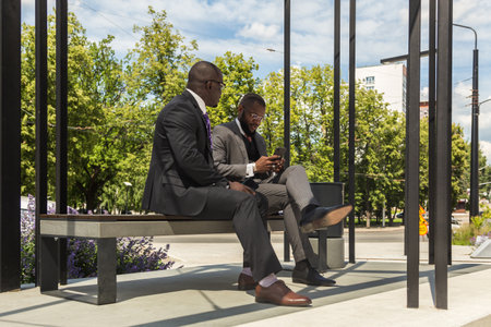 Two Two dark-skinned businessmen in suits communicate. Friends and business partners are sitting on a bench in the city against the backdrop of buildingsの写真素材
