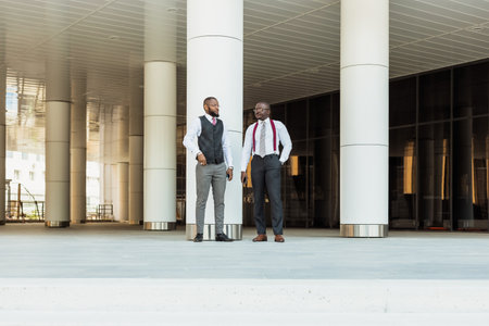 Portrait of two black business partners walking and talking in front of a skyscraper exterior. Friendly meeting outdoorsの写真素材