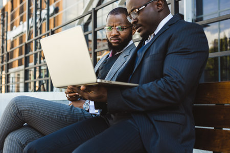 Business meeting of friends outdoors. Two dark-skinned men in suits are sitting on a bench near a city building with a laptop and talking. Partnership and corporate relationsの写真素材