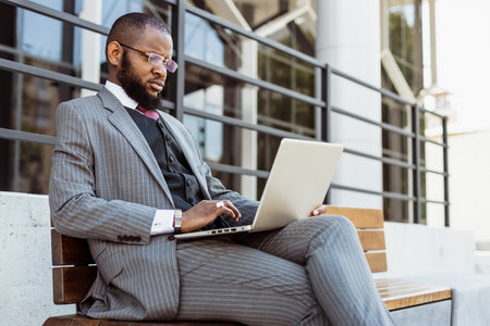 A dark-skinned man in a business suit is using a laptop outdoors. Digital education and remote workの写真素材