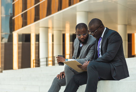 Friendly meeting of business partners outdoors. Two dark-skinned men in suits sit on the steps of a city building with a notebook and have a conversation. Working break. Support and collaborationの写真素材