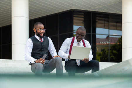 Business meeting of partners outdoors. Two dark-skinned men in suits sit on the steps of a city building with a notebook and a laptop and have a conversation. Friendly support and collaborationの写真素材
